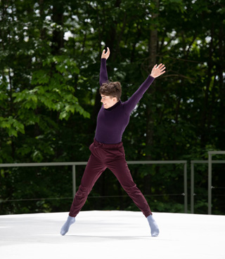 A nonbinary white dancer leaps sideways on an outdoor stage, with their arms flung so that their body makes a big X. They wear a purple turtleneck, maroon pants, and blue socks.