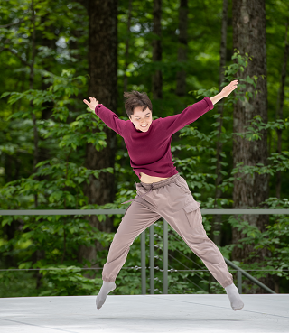 A dancer on an outdoor stage mid-jump. They are wearing a red shirt, tan pants, and grey socks.