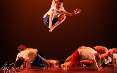 A group of boy dancers on the floor on their hands and feet in a plank position with their feet facing each other to make a circle. There is a boy jumping up in the center of them with his legs tucked and arms extended front.