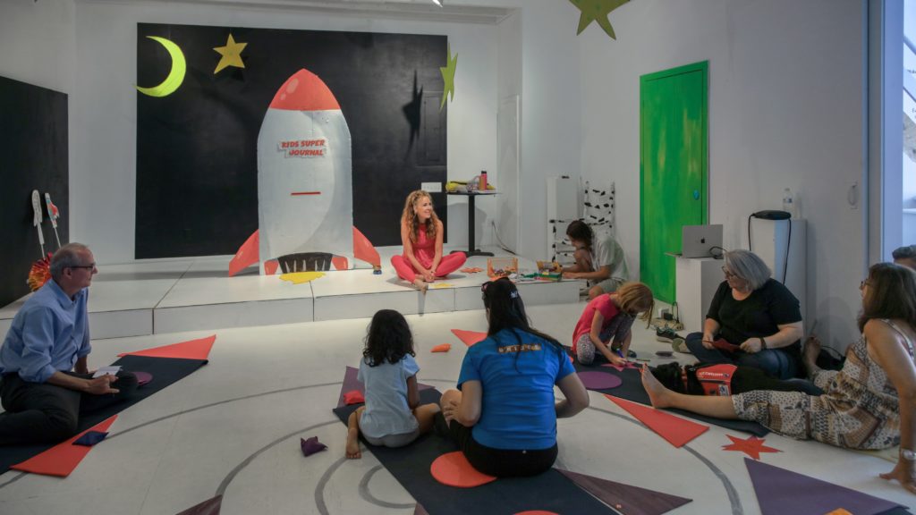 Elizabeth Heller sits in a bright room with colorful space decorations. A group of adults and kids sit around with craft supplies.