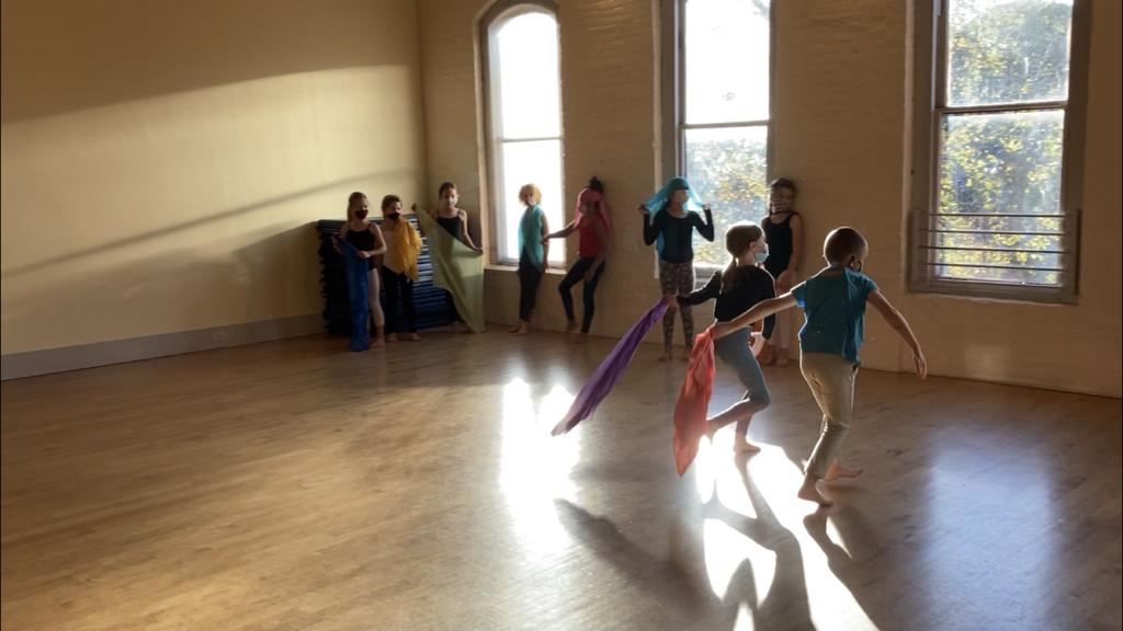 A group of young dancers in a studio carry colorful scarves. Two dancers are in motion running toward a wall with windows while the other dancers line up in the corner and watch.