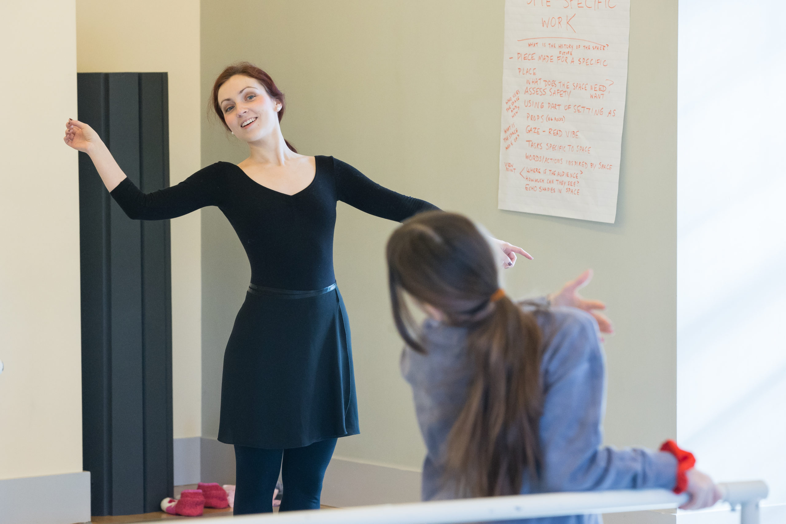 A ballet teacher gestures and leans to demonstrate.