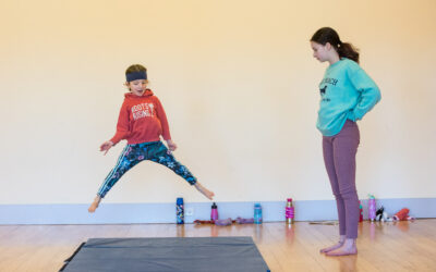 A young dancer jumps in the middle splits while an older dancer watches.