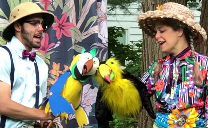 Two puppeteers hold parrot and toucan puppets in an outdoor space.