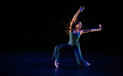 A dancer wearing a blue jumpsuit reaches as she falls to the floor.