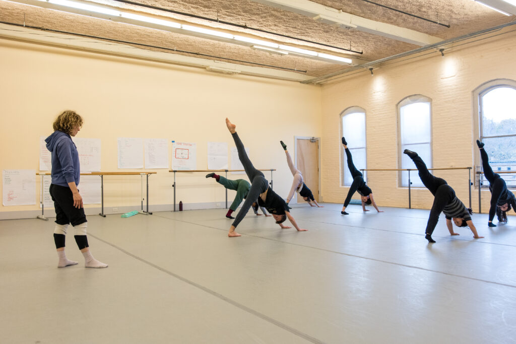 A teacher looks on as a group of students do a three-legged downward dog in a dance studio.