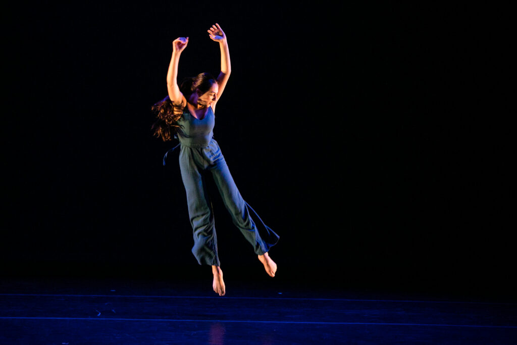 A dancer in a blue jumpsuit jumps, looking down, with her hands thrown above her head.
