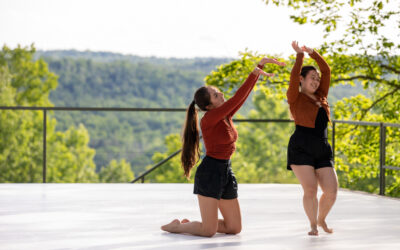 Two dancers on an outdoor stage wearing burnt orange and black shorts. One dancer kneels and raises her arms toward the other while the other dancer completes a turn. Both are smiling.
