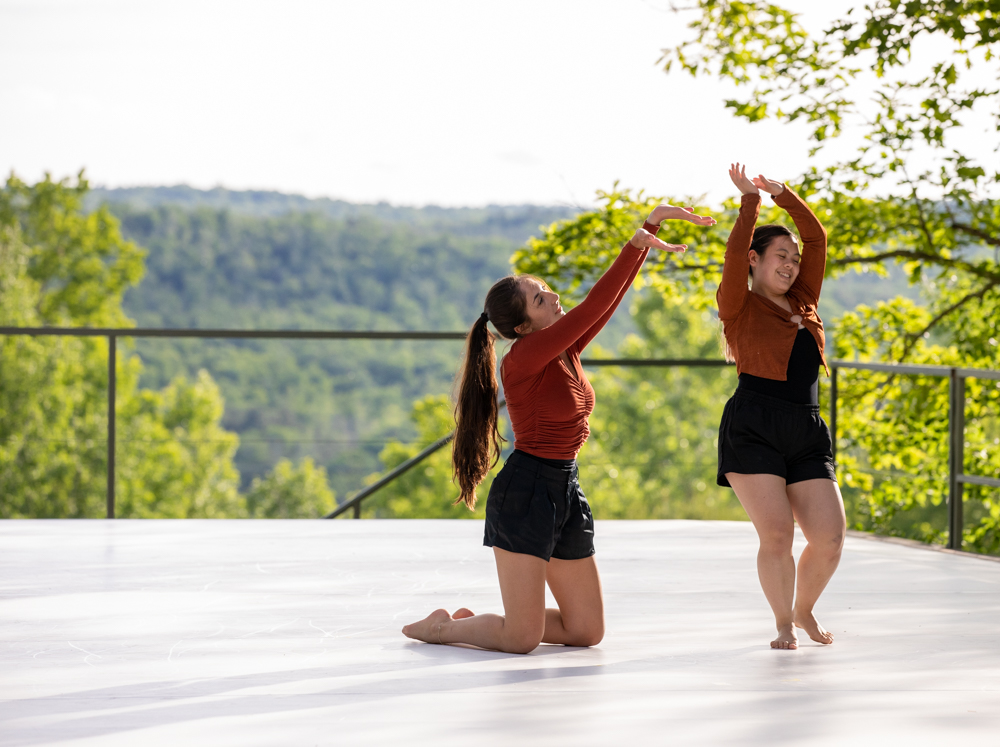 Two dancers on an outdoor stage wearing burnt orange and black shorts. One dancer kneels and raises her arms toward the other while the other dancer completes a turn. Both are smiling.