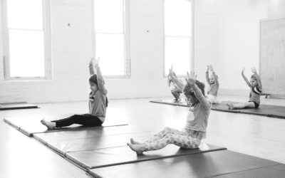 A black and white photo of young dancers stretching on mats in a studio. They are sitting and reach their arms overhead.