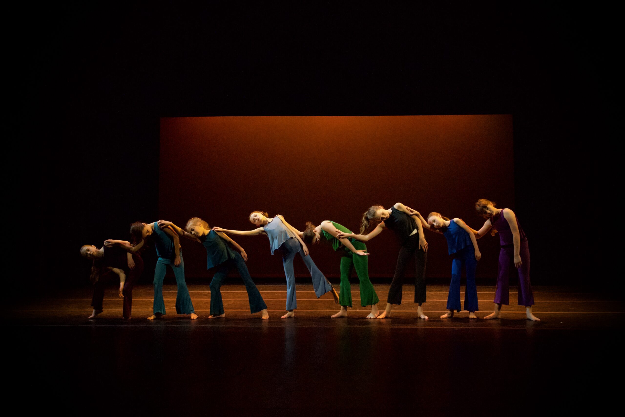 A line of dancers wearing blues, greens, and purples leaning on each other. They stand on a stage with orange lighting.