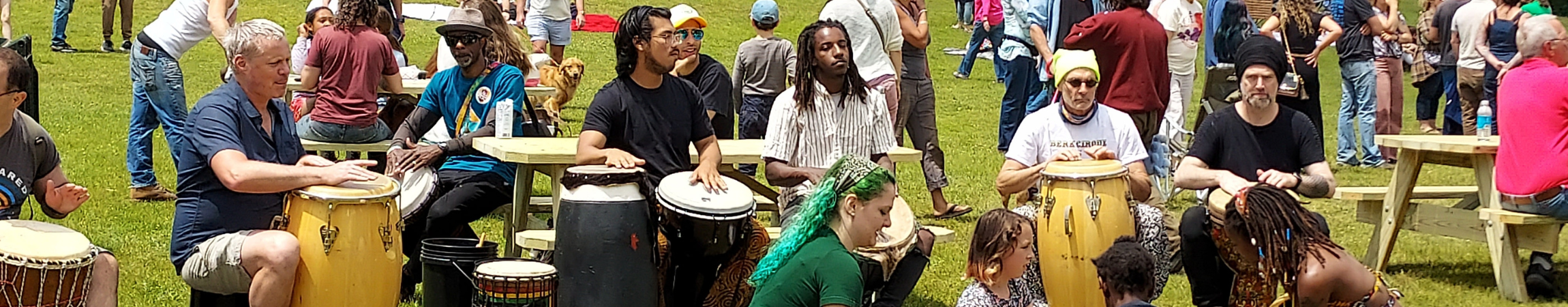 A group of drummers play outside while people dance and mill about.
