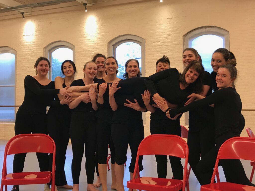 A group of young dancers hold up Diane Pearlman in a studio. They are all wearing black and there are red chairs in front of their legs.